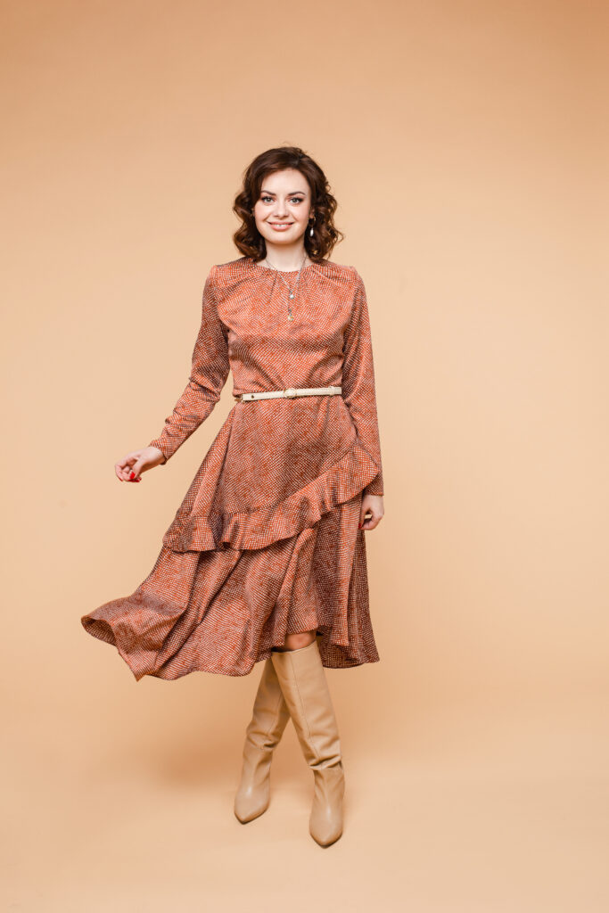 Full length stock photo of a beautiful wavy-haired brunette Caucasian woman wearing lovely coral dress with beige belt on waist and high beige leather boots. Her skirt is in motion. Smiling at camera. Isolated.