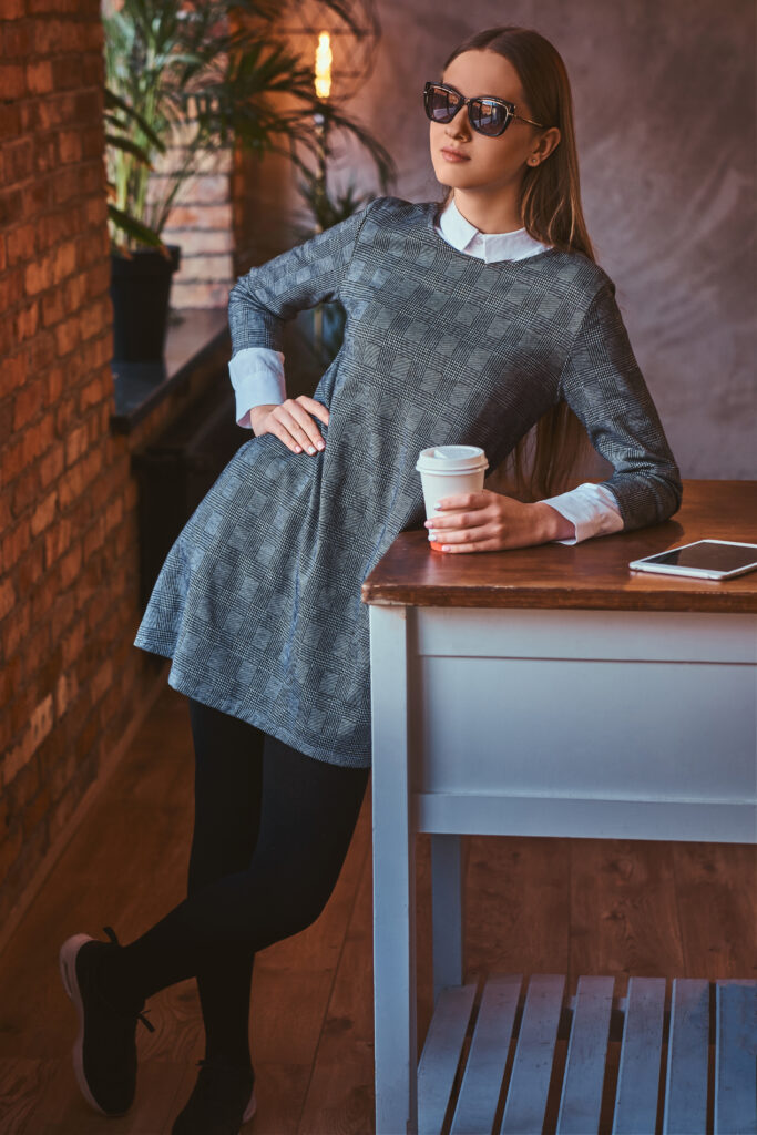 Portrait of a smiling young girl on sunglasses dressed in an elegant gray dress holds a cup of takeaway coffee and tablet while leaning on the table in a room with loft interior.