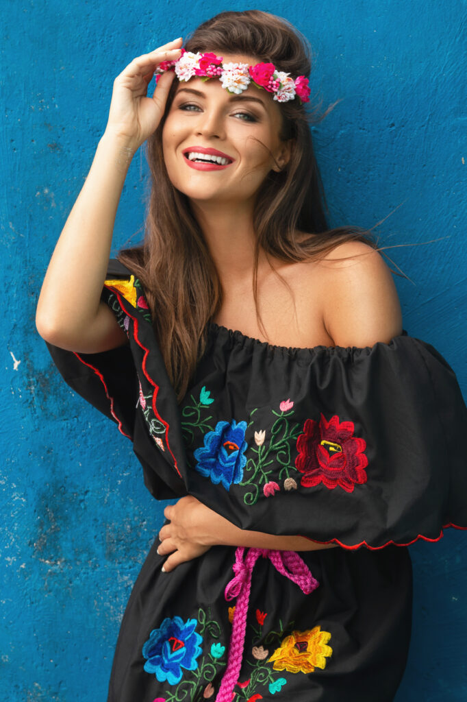 Young beautiful woman wearing traditional mexican dress is posing beside blue concrete wall